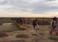 Fila de peregrinos wixáritari con vestimenta tradicional bordada y sombreros adornados caminando por un terreno semiárido, bajo un cielo nublado al atardecer.