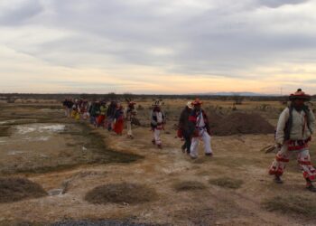 Fila de peregrinos wixáritari con vestimenta tradicional bordada y sombreros adornados caminando por un terreno semiárido, bajo un cielo nublado al atardecer.