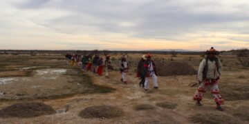 Fila de peregrinos wixáritari con vestimenta tradicional bordada y sombreros adornados caminando por un terreno semiárido, bajo un cielo nublado al atardecer.