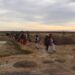 Fila de peregrinos wixáritari con vestimenta tradicional bordada y sombreros adornados caminando por un terreno semiárido, bajo un cielo nublado al atardecer.