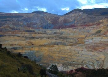Gran tajo abierto con bancos escalonados de tonos amarillos, grises y rojizos, rodeado de laderas con vegetación y bajo un cielo nublado al atardecer.
