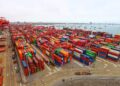 Vista aérea de un terminal portuario con cientos de contenedores de colores apilados, grúas pórtico rojas y el mar con embarcaciones al fondo.