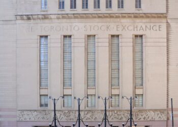 Fachada de piedra clara de estilo art déco con la inscripción The Toronto Stock Exchange, ventanales verticales y faroles frente a la calle, entre dos torres de vidrio.