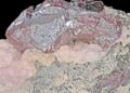 Primer plano de un mineral de plata de tonos rojizos y metálicos sobre una base de roca rosada y gris, con fondo negro.