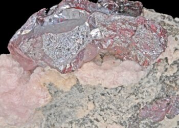 Primer plano de un mineral de plata de tonos rojizos y metálicos sobre una base de roca rosada y gris, con fondo negro.