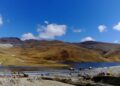 Paisaje altoandino con una laguna y cerros ocres bajo cielo azul; en primer plano, una plataforma de tierra con maquinaria y vehículos mineros.