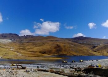 Paisaje altoandino con una laguna y cerros ocres bajo cielo azul; en primer plano, una plataforma de tierra con maquinaria y vehículos mineros.