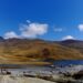 Paisaje altoandino con una laguna y cerros ocres bajo cielo azul; en primer plano, una plataforma de tierra con maquinaria y vehículos mineros.
