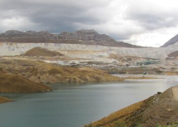 Mina Antamina en los Andes de Áncash vista desde el sureste, con su presa de relaves de agua verdosa en primer plano, instalaciones industriales al fondo y cerros bajo un cielo nublado.