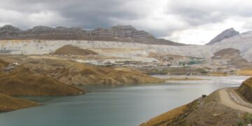 Mina Antamina en los Andes de Áncash vista desde el sureste, con su presa de relaves de agua verdosa en primer plano, instalaciones industriales al fondo y cerros bajo un cielo nublado.