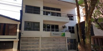 Fachada de un edificio blanco de tres pisos con el letrero verde de la Agencia Nacional de Minería sobre la entrada, junto a un árbol, en una calle de Medellín.
