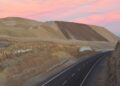Camino asfaltado que atraviesa un corte de terreno junto a grandes depósitos de material de la mina de cobre Mantos Blancos, en Antofagasta, bajo un cielo rosado al atardecer.
