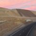 Camino asfaltado que atraviesa un corte de terreno junto a grandes depósitos de material de la mina de cobre Mantos Blancos, en Antofagasta, bajo un cielo rosado al atardecer.