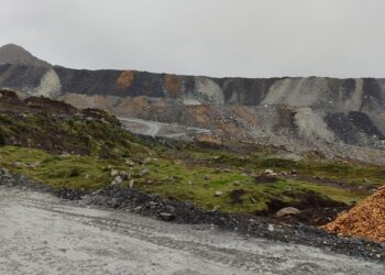 Camino de tierra mojado frente a grandes depósitos escalonados de material de color gris y ocre en la mina Antamina, con pasto verde en primer plano y cielo nublado.