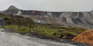 Camino de tierra mojado frente a grandes depósitos escalonados de material de color gris y ocre en la mina Antamina, con pasto verde en primer plano y cielo nublado.